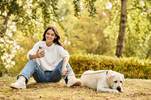 A beautiful young woman relaxes in the park, checking her phone beside her dog on a warm autumn day.