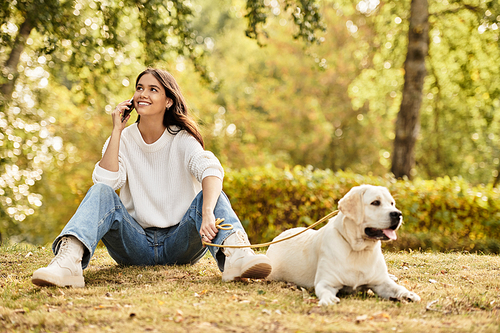 A young woman dressed in cozy autumn attire relaxes outdoors, chatting happily on her phone.