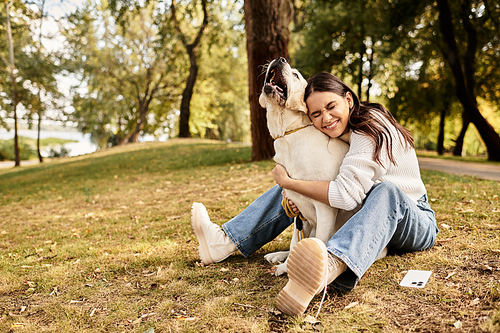 A delighted woman embraces her dog while sitting on the grass in a vibrant autumn park.