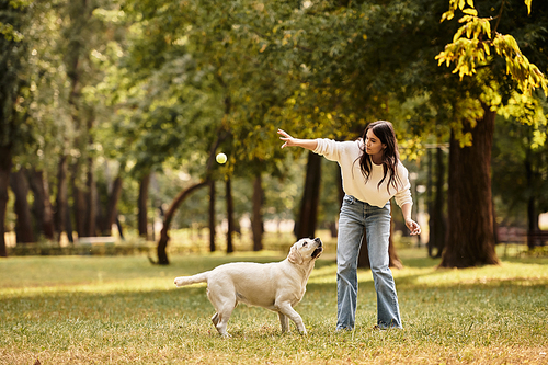 A young woman in cozy autumn clothes happily tosses a ball for her dog in a park.