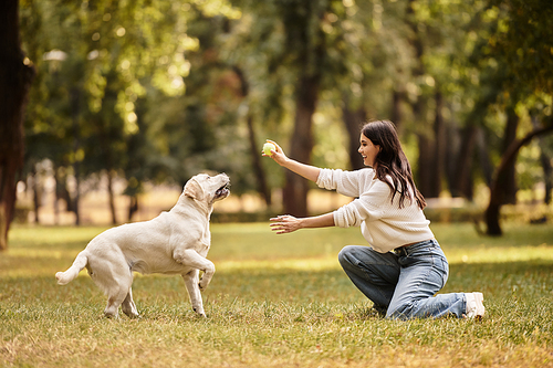 A beautiful woman in cozy attire kneels in the park, tossing a ball to her energetic dog.