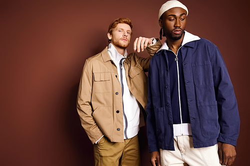 Two stylish multicultural men standing together in front of a brown wall.