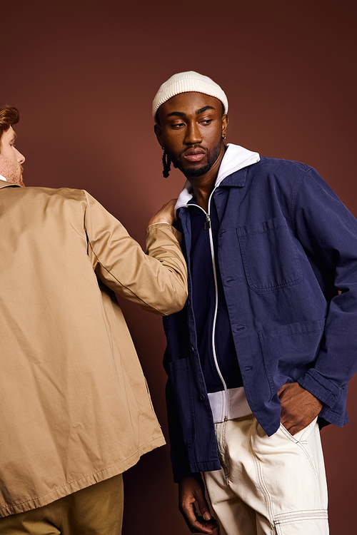 Two multicultural men in stylish attire stand together against a brown background.