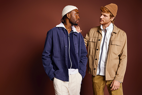 Two stylish multicultural men pose elegantly in front of a brown wall.