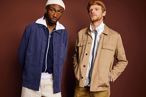 Two handsome men with stylish flair stand side by side in front of a wall.