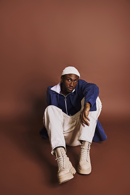 Handsome young African American man in stylish outfit sitting cross-legged on the ground.