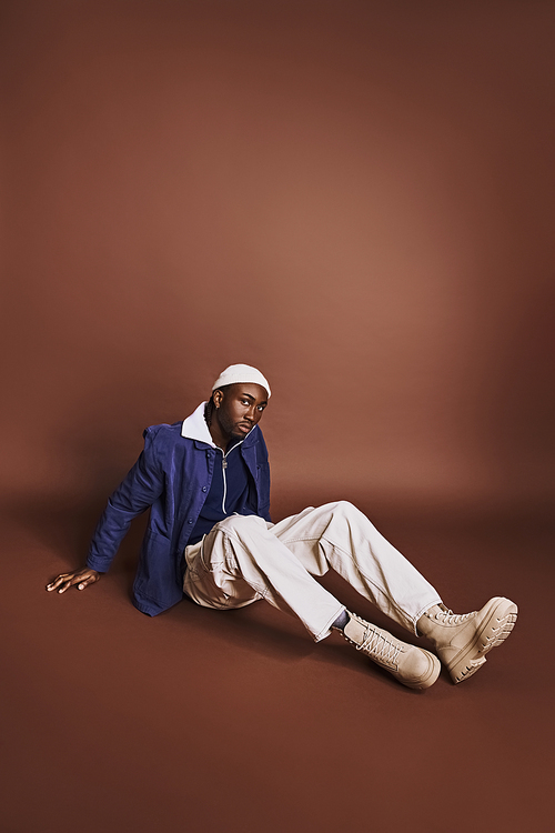 Handsome African American man sitting on ground in blue jacket and white pants.