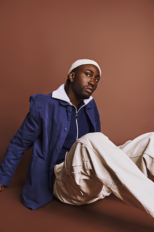 Stylish African American man sitting on ground in blue jacket and white pants.