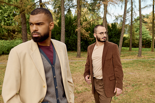 Two fashionable men enjoy a leisurely walk in an autumn field amidst colorful foliage and tall trees.