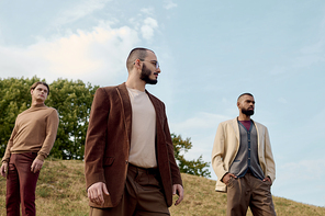 Three handsome men dressed in autumn attire pose confidently in a tranquil natural setting.