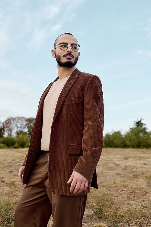 A handsome man poses gracefully in a stylish autumn outfit amidst a beautiful natural landscape.