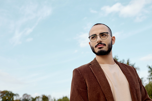 A handsome man dressed in autumn attire stands confidently in a serene natural setting, enjoying the day.