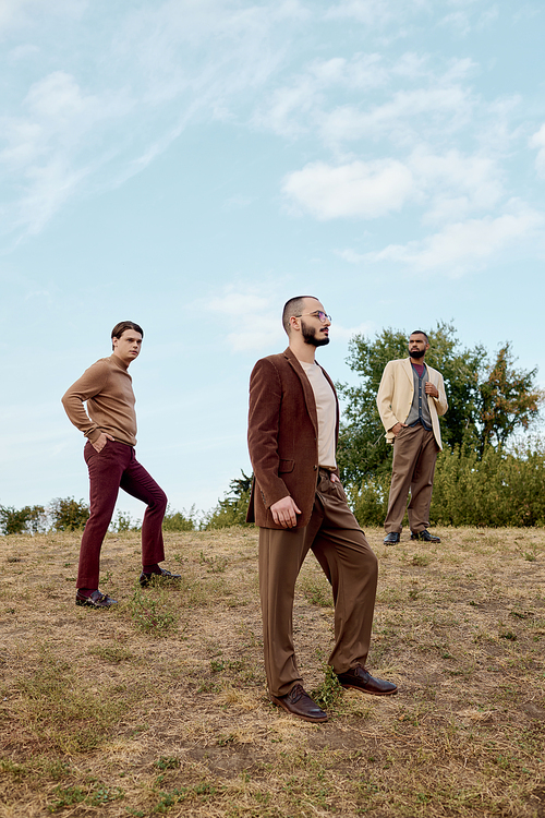 Three confident men pose in elegant autumn outfits against a serene natural backdrop, celebrating style.