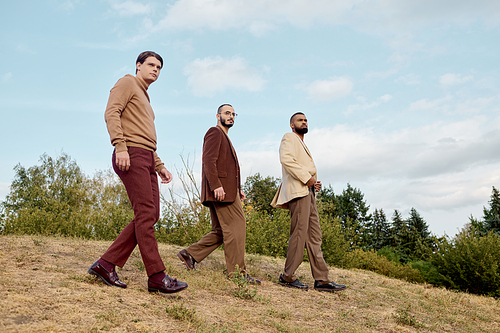 Three handsome men dressed in autumn attire stroll confidently through a beautiful field landscape.