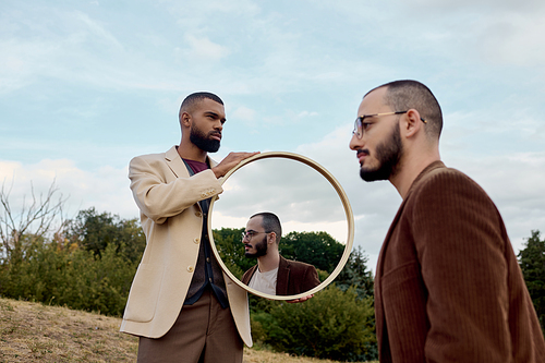 Two handsome men dressed in fashionable autumn attire pose thoughtfully against a natural landscape.