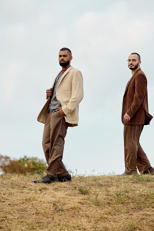 Two handsome men in stylish autumn attire pose confidently in a picturesque field at sunset.