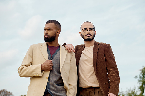 Two handsome men showcase their fashionable autumn outfits, posing confidently in a tranquil field setting.