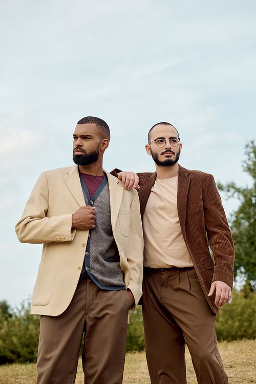 Two handsome men dressed in fashionable fall attire stand confidently in a serene outdoor landscape.