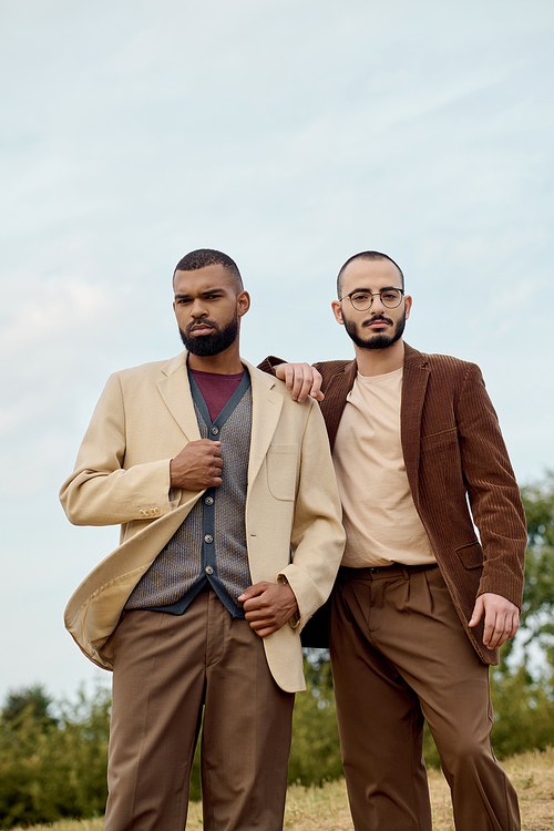 Two handsome men dressed in fashionable autumn attire pose confidently in a picturesque natural setting.