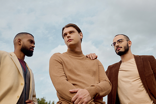 Three handsome men pose confidently in a tranquil field, displaying trendy autumn attire and camaraderie.