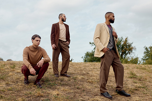 Three handsome men in trendy autumn attire pose confidently in a tranquil field under cloudy skies.