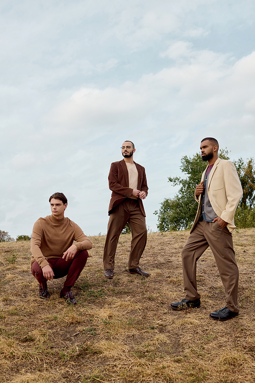 Three handsome men stand in a serene autumn landscape, dressed in fashionable outfits that blend elegance and nature.