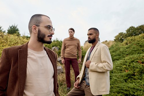 Three handsome men model trendy autumn outfits in a picturesque field surrounded by nature.