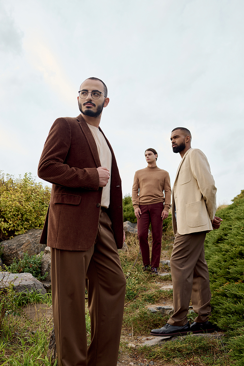 Three handsome men pose confidently in autumn attire, surrounded by vibrant nature under a cloudy sky.