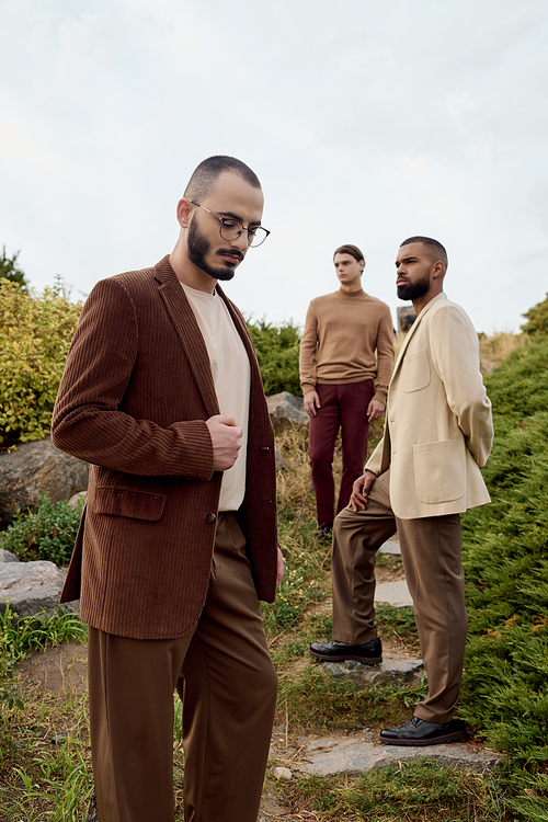 Three handsome men in fashionable autumn outfits pose confidently amidst a picturesque field at dusk.