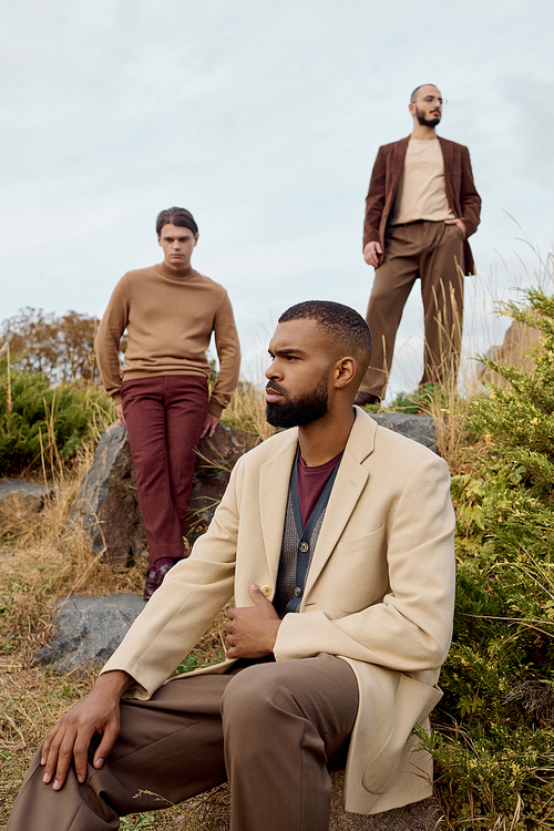 Three handsome men in fashionable autumn outfits pose thoughtfully amid a beautiful natural landscape.