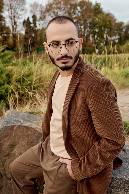 A handsome man in fashionable autumn clothing stands proudly in a tranquil field surrounded by nature.