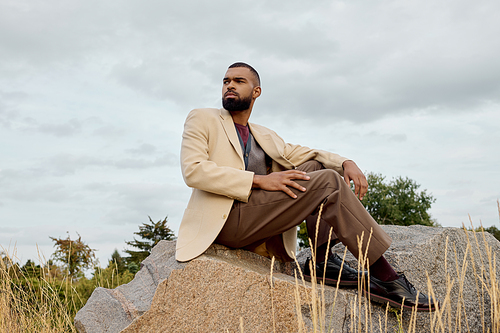 A handsome man dressed in fashionable autumn attire poses thoughtfully in a tranquil field, surrounded by nature.