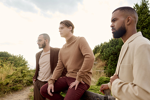 Three handsome men dressed in autumn fashion pose confidently in a tranquil outdoor landscape.