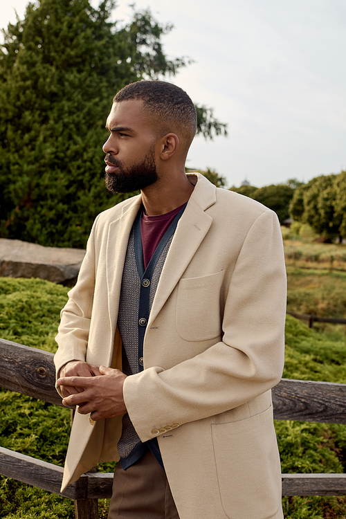 A well-dressed man poses thoughtfully among vibrant autumn foliage in a tranquil outdoor setting.