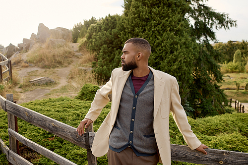 A handsome man in chic autumn attire stands gracefully against a natural backdrop, embracing the season beauty.