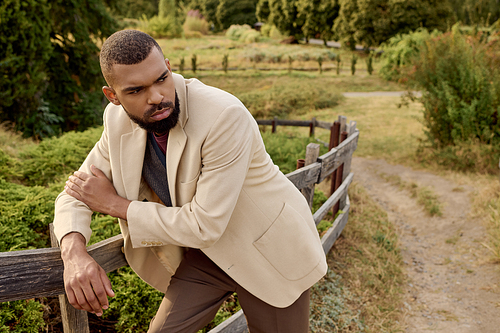 A handsome man leans against a wooden fence, dressed in chic autumn attire surrounded by nature.