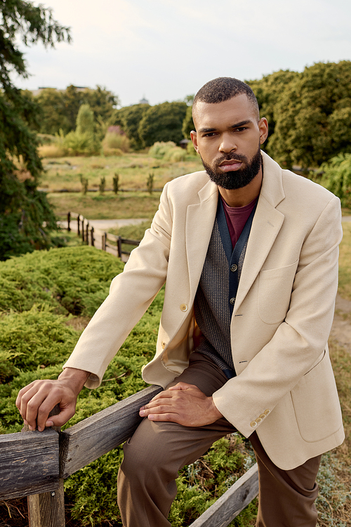 A dapper man stands confidently on a wooden fence, showcasing autumn fashion in a tranquil natural setting.