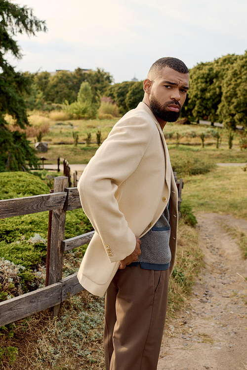 A stylish man poses confidently in a scenic field, surrounded by the warm hues of autumn foliage.