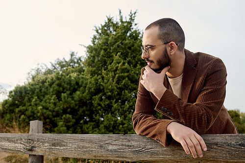 A handsome man in trendy autumn attire relaxes against a wooden fence in a tranquil field setting.