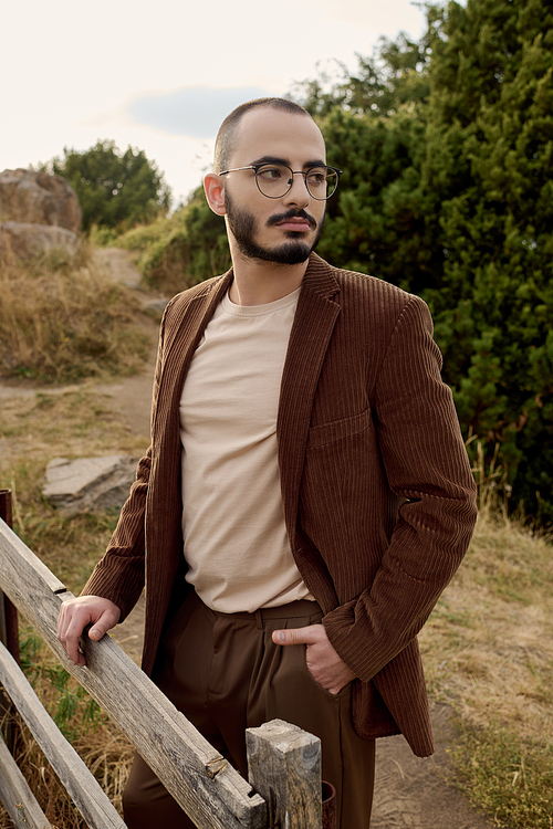 A handsome man dressed in autumn attire stands confidently in a picturesque field surrounded by nature.