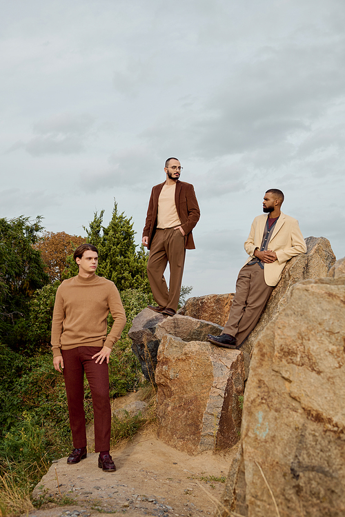 Three stylish men pose confidently outdoors, showcasing fashionable autumn outfits in a vibrant setting.