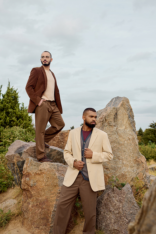 Two handsome men dressed in stylish autumn attire pose confidently amongst natural rocks in a tranquil field.