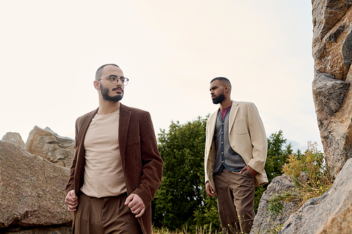 Two handsome men in chic autumn outfits stroll through a serene natural landscape, exuding confidence.