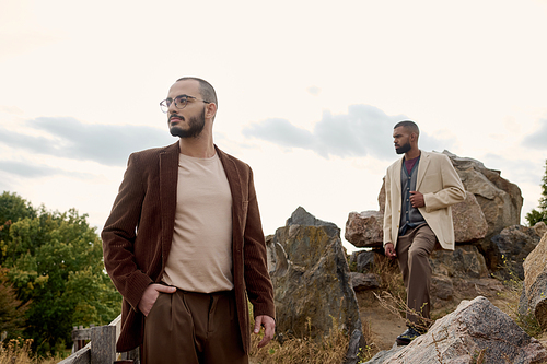 Two handsome men dressed in stylish autumn attire stand confidently in a picturesque outdoor field.