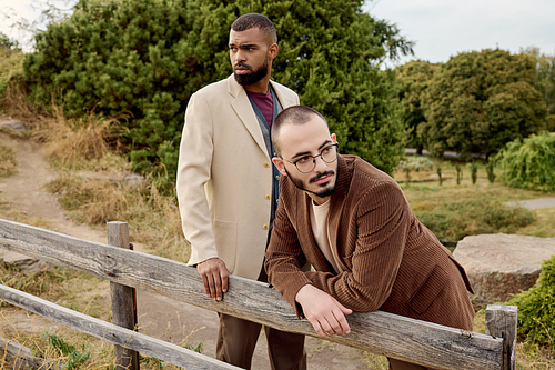 Two handsome men dressed in stylish autumnal outfits strike a pose amidst the tranquil beauty of nature.