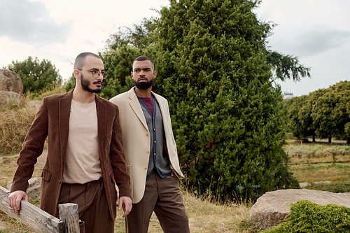 Two handsome men donning fashionable autumn attire pose confidently amidst a picturesque landscape.