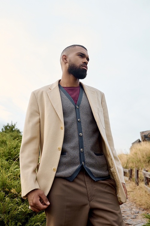 A handsome man dressed in autumn attire stands confidently in a tranquil field surrounded by nature.