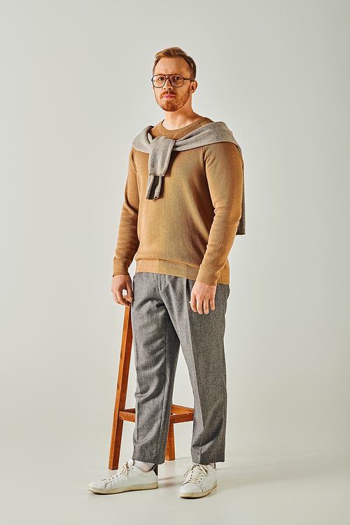 A stylish man poses on a stool with a sweater elegantly draped around his neck.