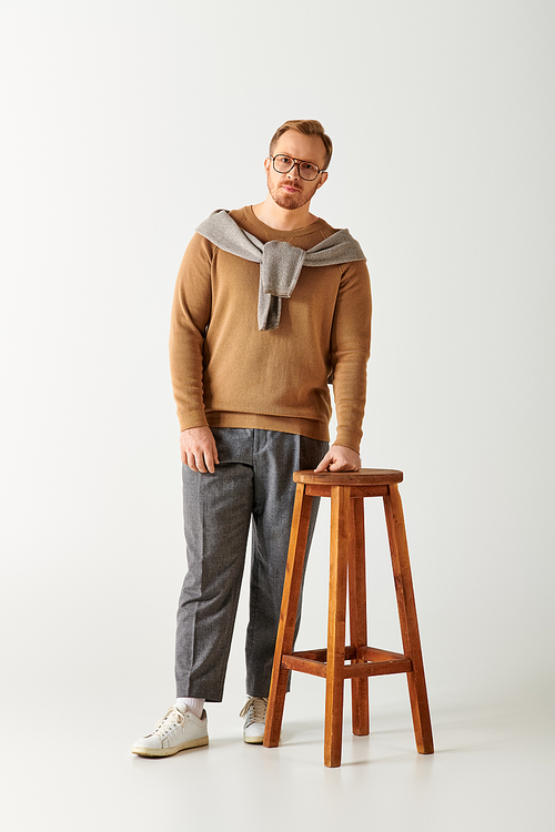 Handsome man in stylish attire posing next to a wooden stool.