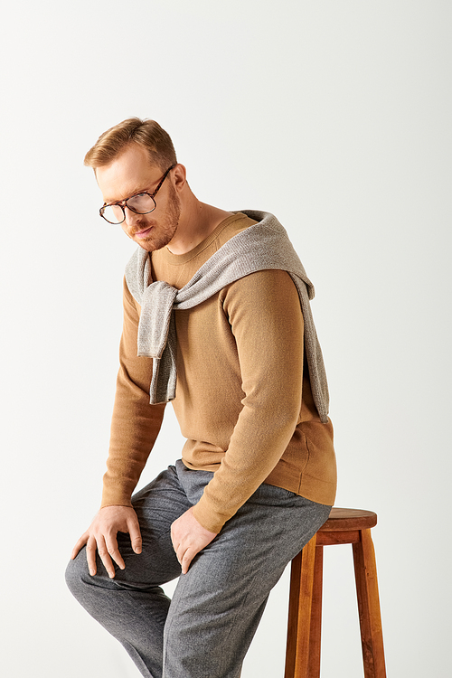 A handsome man in stylish attire actively poses while sitting on top of a wooden stool.